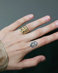 A close-up of a hand wearing three gold rings, with a small black geometric tattoo on the ring finger. The background is a dark, neutral gray.