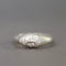 A sterling silver ring featuring a sculpted face with closed eyes and a small clear gemstone on the cheek, set against a neutral gray background.