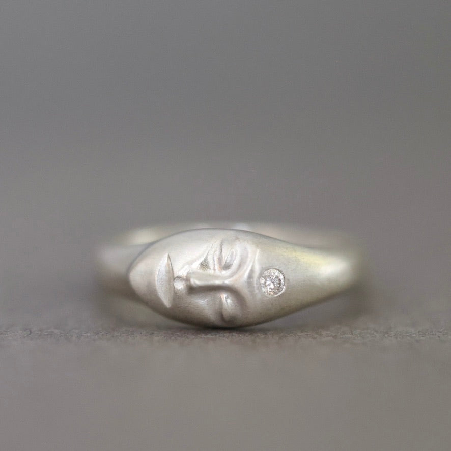 A sterling silver ring featuring a sculpted face with closed eyes and a small clear gemstone on the cheek, set against a neutral gray background.