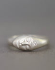 A sterling silver ring featuring a sculpted face with closed eyes and a small clear gemstone on the cheek, set against a neutral gray background.