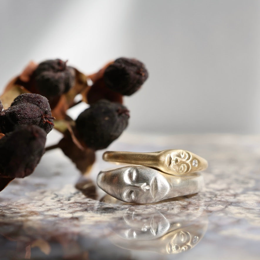 Two face-shaped rings, one gold and one silver, displayed on a reflective surface with dried dark berries in the background.
