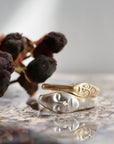Two face-shaped rings, one gold and one silver, displayed on a reflective surface with dried dark berries in the background.