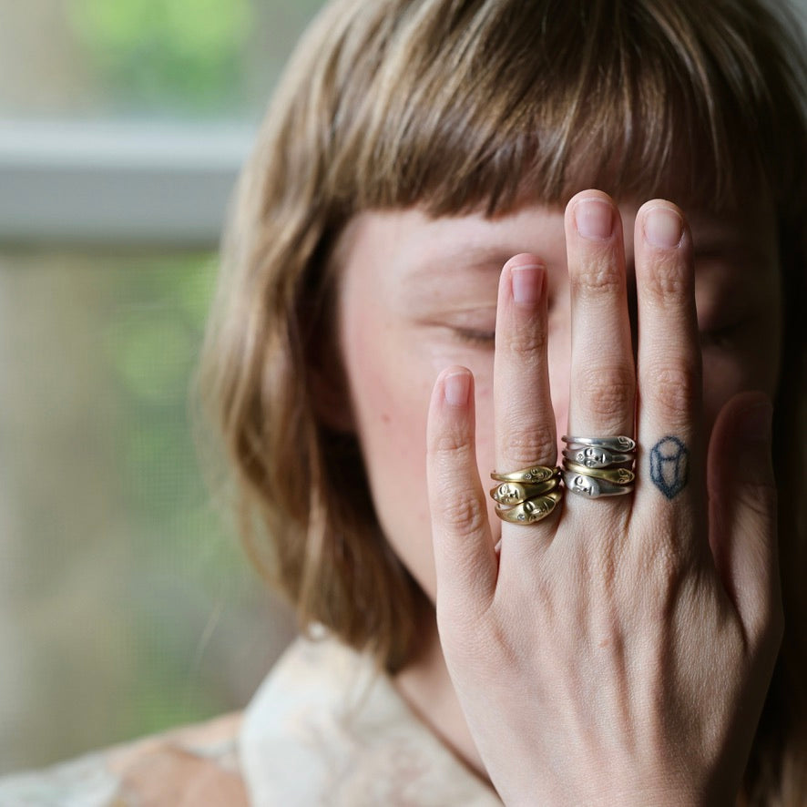 A close-up lifestyle shot of a person with short brown hair covering their face with their hand, showcasing several rings on their fingers. The rings include a textured gold ring, a silver ring with a geometric design, and a silver ring with a white logo.