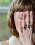 A close-up lifestyle shot of a person with short brown hair covering their face with their hand, showcasing several rings on their fingers. The rings include a textured gold ring, a silver ring with a geometric design, and a silver ring with a white logo.