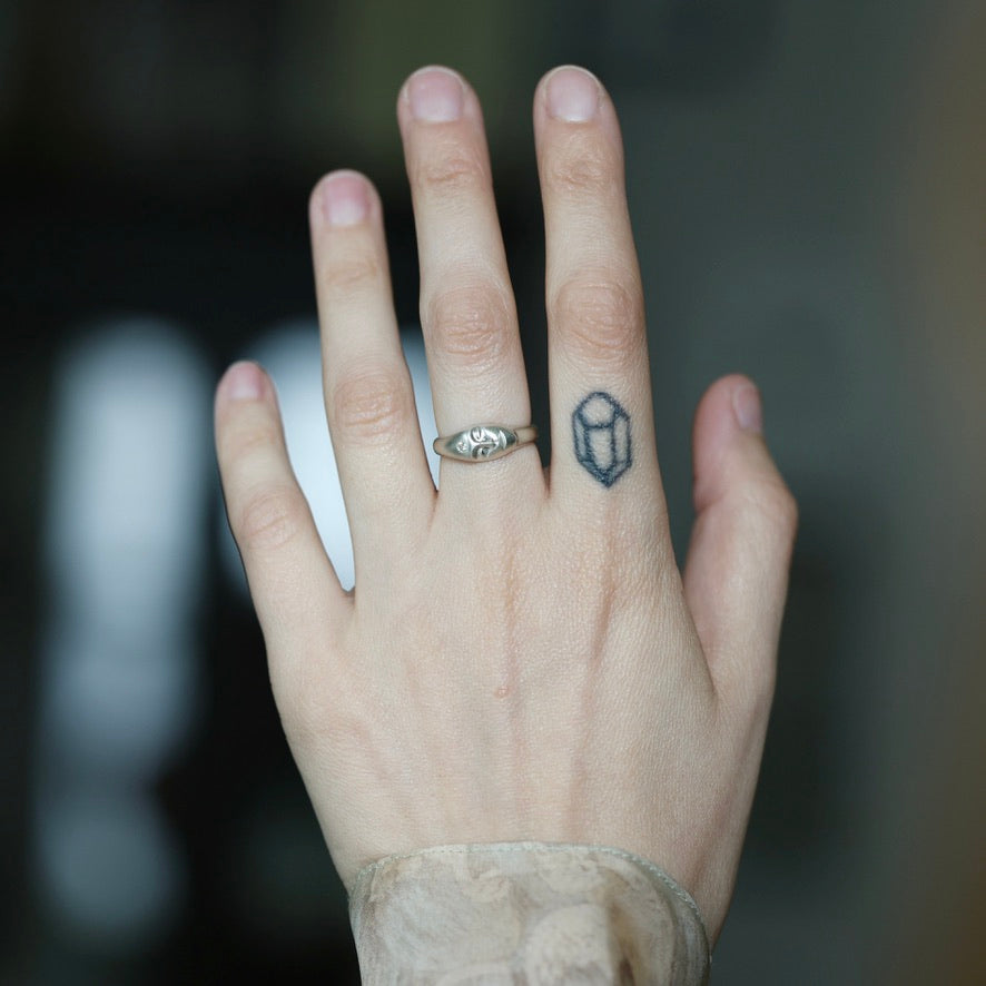 A close-up of a person's hand showing two rings: a silver band ring with a small engraved design on the ring finger, and a small dark geometric line-art ring on the middle finger.
