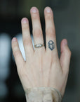 A close-up of a person's hand showing two rings: a silver band ring with a small engraved design on the ring finger, and a small dark geometric line-art ring on the middle finger.