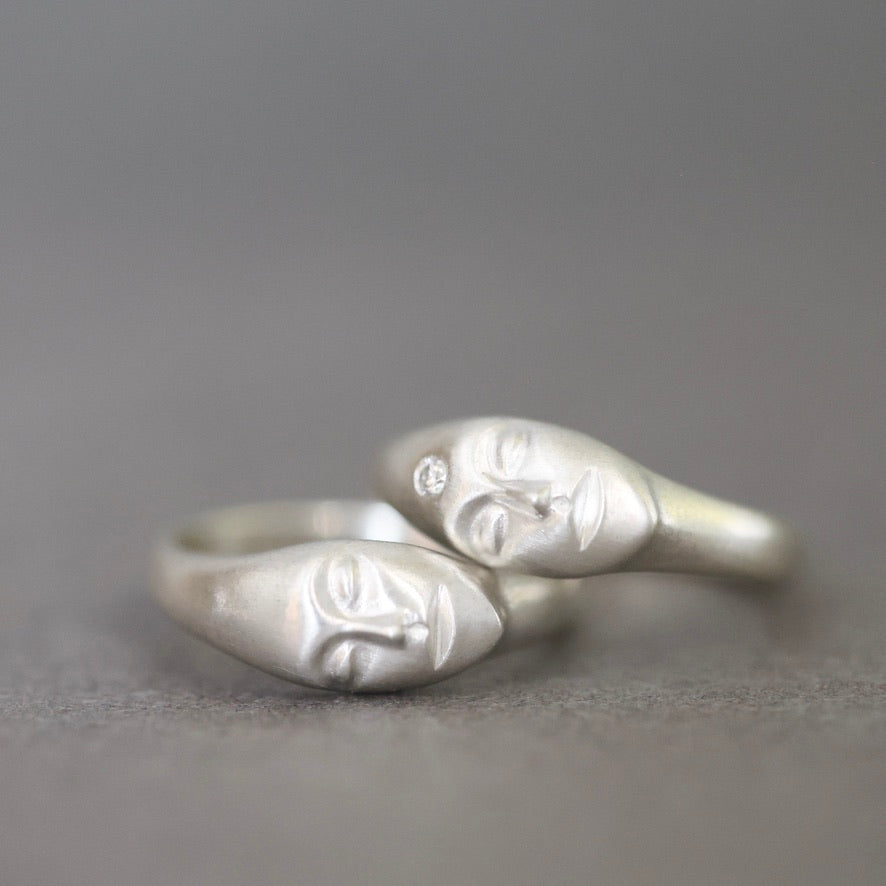 Two silver rings shaped like stylized faces, one with a small clear stone, photographed on a smooth gray surface.