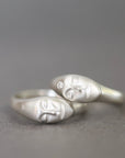 Two silver rings shaped like stylized faces, one with a small clear stone, photographed on a smooth gray surface.