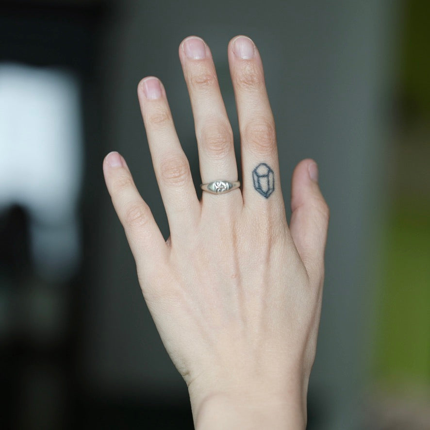 A close-up of a person's hand showing a silver ring with a small geometric cube design on the ring finger, set against a blurred dark background.