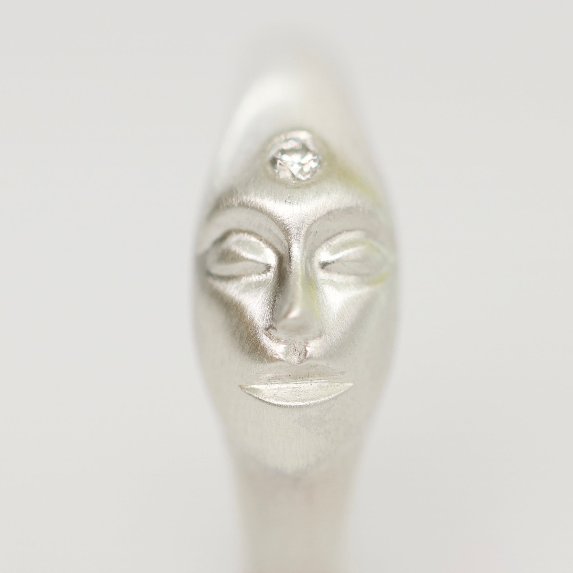 A close-up of a silver-toned sculptural face pendant or charm with closed eyes and a small clear gemstone set above the forehead, shown against a plain light gray background.
