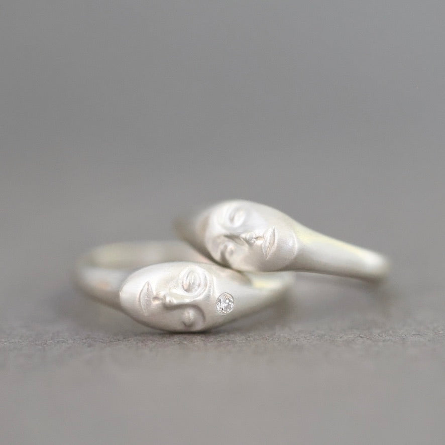 A pair of silver rings shaped like stylized faces, with one ring featuring a small clear gemstone, set against a soft gray background.