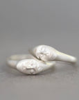 A pair of silver rings shaped like stylized faces, with one ring featuring a small clear gemstone, set against a soft gray background.