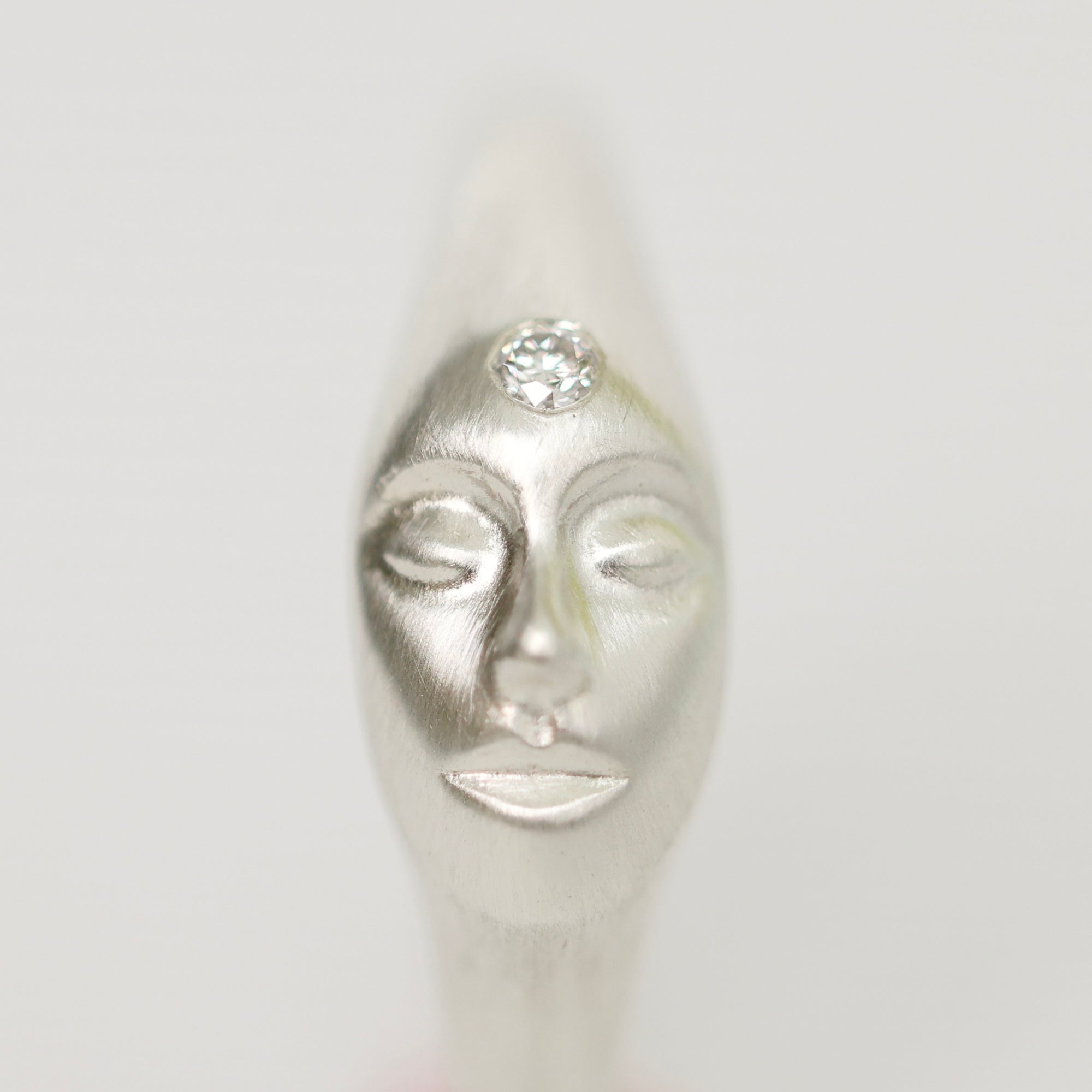 A silver sculpted ring shaped like a stylized face with closed eyes and a clear round gemstone set on the forehead, shown against a plain light gray background.