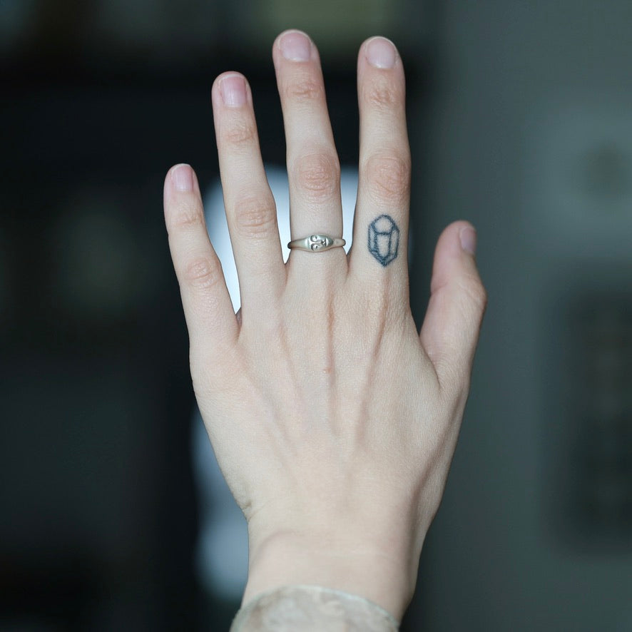 A close-up of a person's hand wearing a silver ring with a small geometric gemstone on the ring finger, with a small tattoo on the side of the finger.