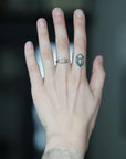 A close-up of a person's hand wearing a silver ring with a small geometric gemstone on the ring finger, with a small tattoo on the side of the finger.