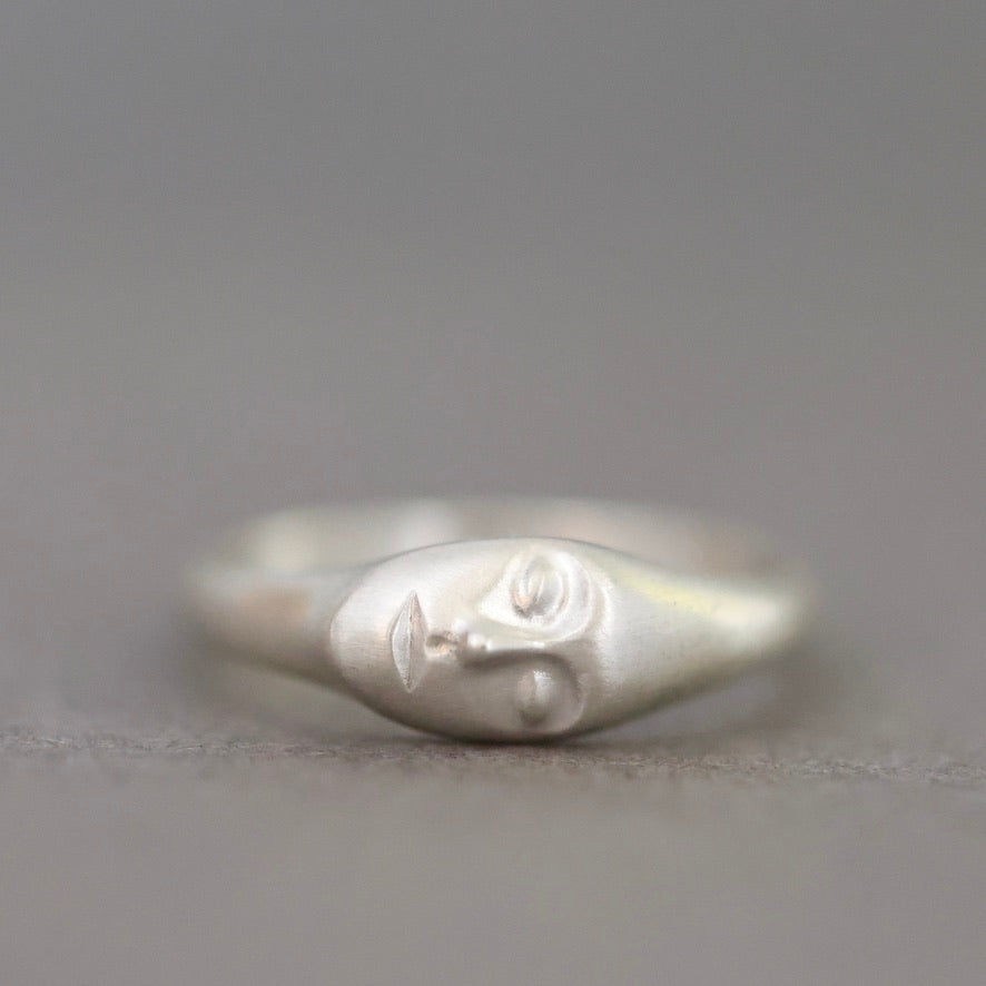 A sterling silver ring featuring a stylized, minimalist face with closed eyes, set against a soft, out-of-focus gray background.