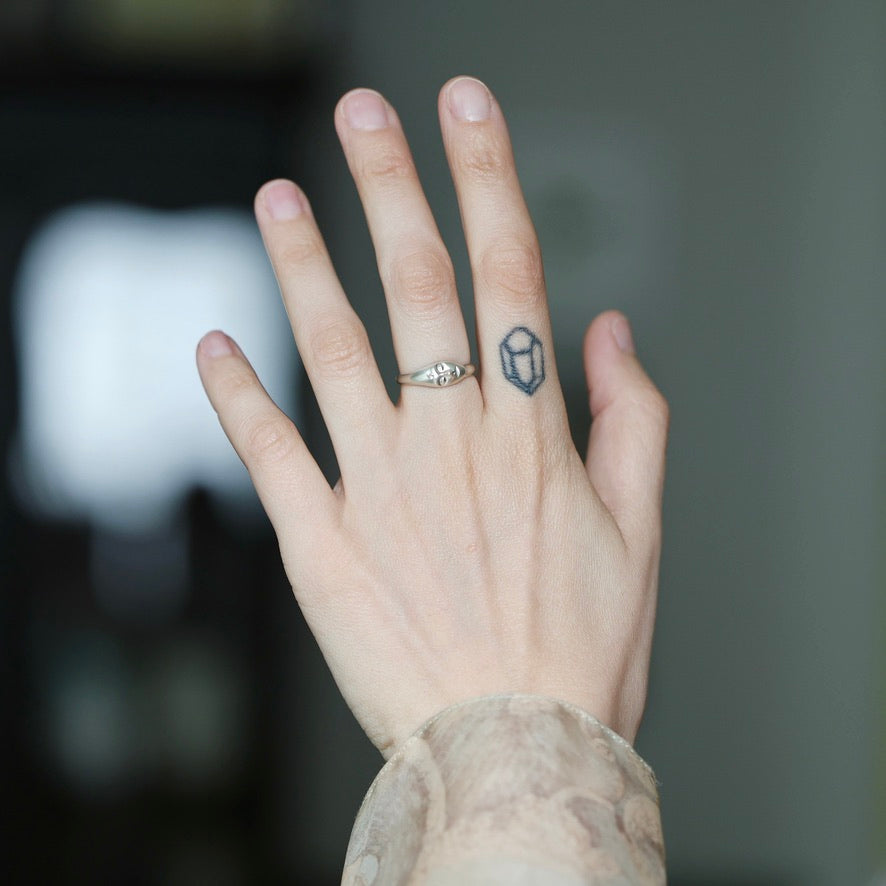 A close-up of a person's hand showing two rings: a silver band ring with a small clear stone on the ring finger, and a small dark geometric tattoo or symbol on the middle finger.