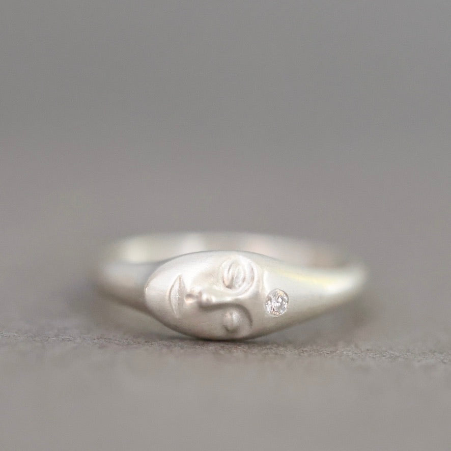 A sterling silver ring shaped like a stylized face with a small clear gemstone, photographed on a neutral gray background.
