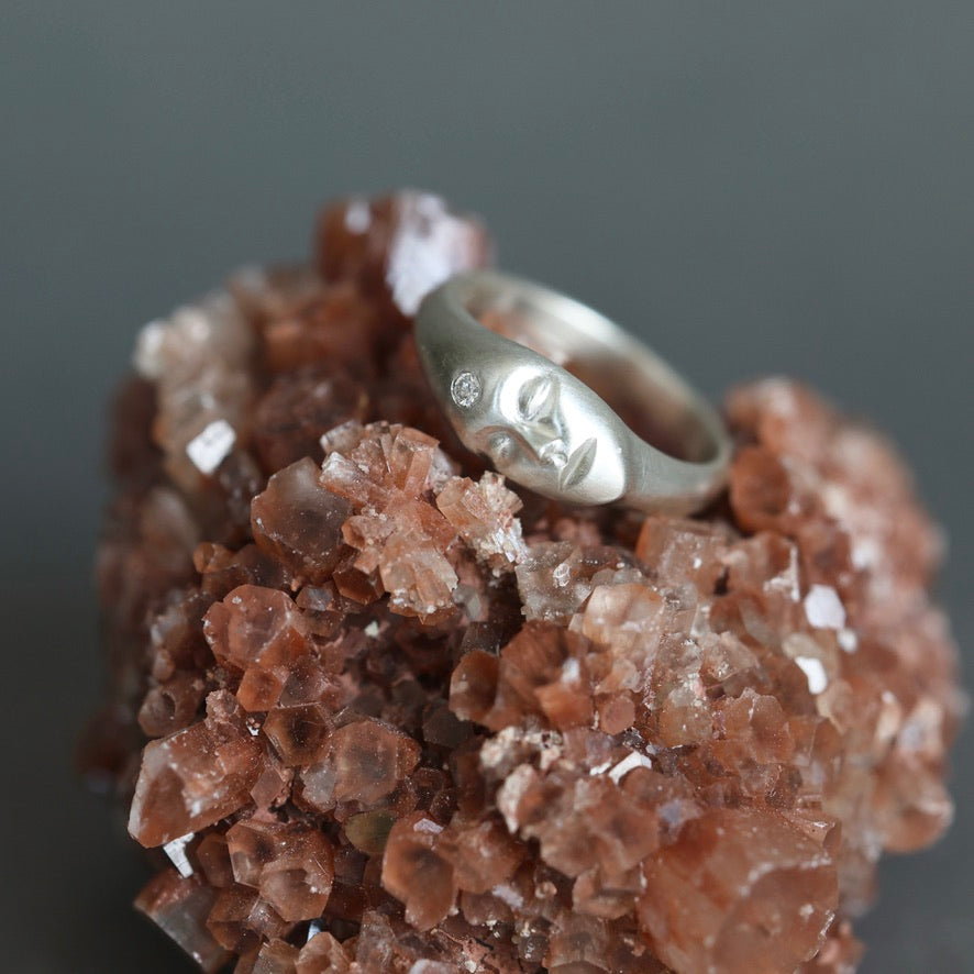 A silver ring with a stylized face engraving and a small clear stone, displayed on a textured brown crystal cluster against a dark gray background.