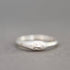 A sterling silver ring featuring a sculpted face with closed eyes and a small round clear gemstone on its cheek, set against a neutral gray background.