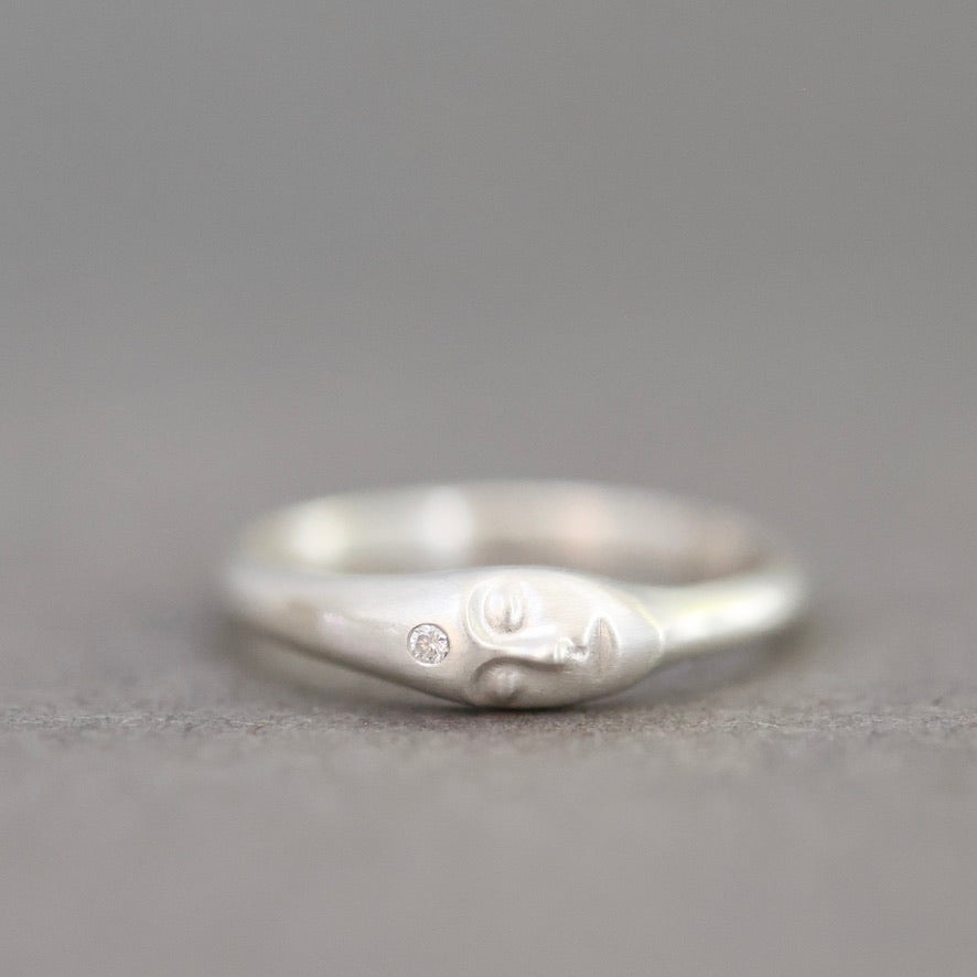 A sterling silver ring featuring a sculpted face with closed eyes and a small round clear gemstone on its cheek, set against a neutral gray background.