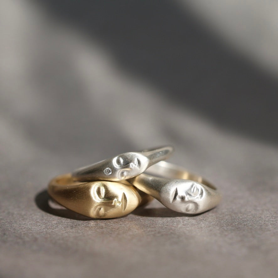 Three sculptural face-shaped rings in polished yellow gold, silver, and white gold, stacked together on a textured gray surface with soft shadows.