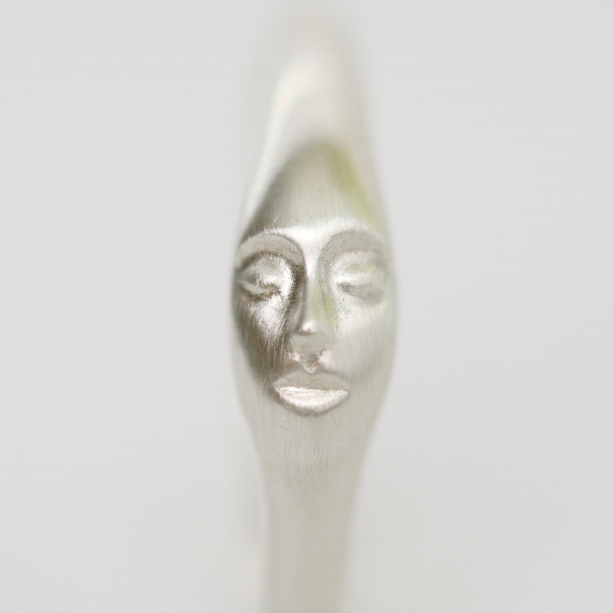A close-up of a metallic silver spoon handle sculpted with a stylized human face, shown against a plain light gray background.