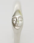 A close-up of a metallic silver spoon handle sculpted with a stylized human face, shown against a plain light gray background.