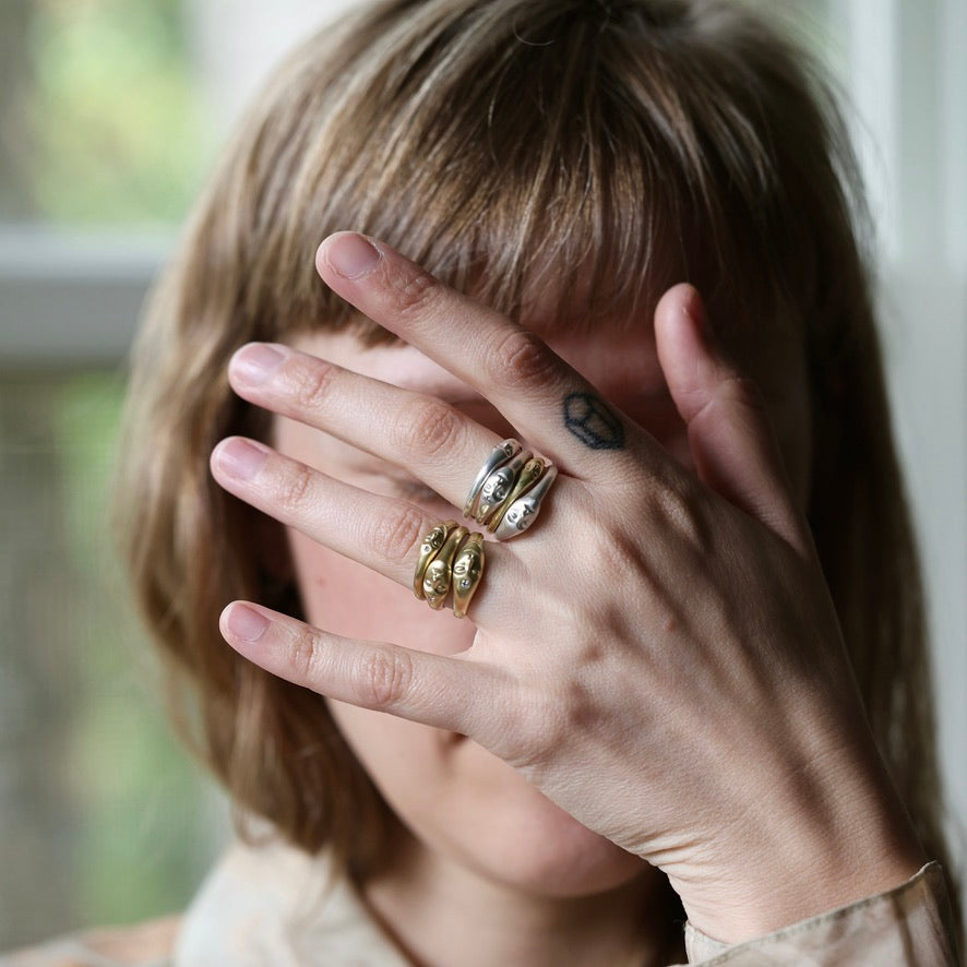 A close-up lifestyle shot of a person with short blonde hair covering their face with their hand, showcasing three gold rings: a textured band, a polished band with a small heart engraving, and a multi-band ring with gold leaf detailing.