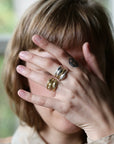 A close-up lifestyle shot of a person with short blonde hair covering their face with their hand, showcasing three gold rings: a textured band, a polished band with a small heart engraving, and a multi-band ring with gold leaf detailing.