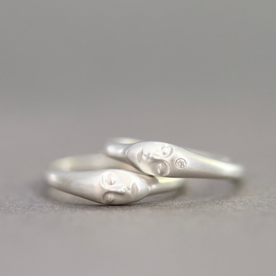 A pair of silver rings shaped like stylized faces, resting on a neutral gray surface.