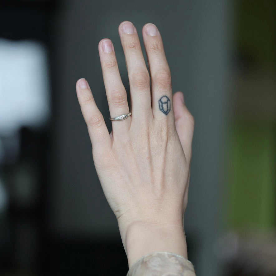 A close-up of a person's hand showing a silver band ring with a small clear gemstone on the ring finger and a small geometric tattoo on the middle finger, set against a blurred dark background.
