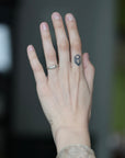 A close-up of a person's hand showing a silver band ring with a small clear gemstone on the ring finger and a small geometric tattoo on the middle finger, set against a blurred dark background.