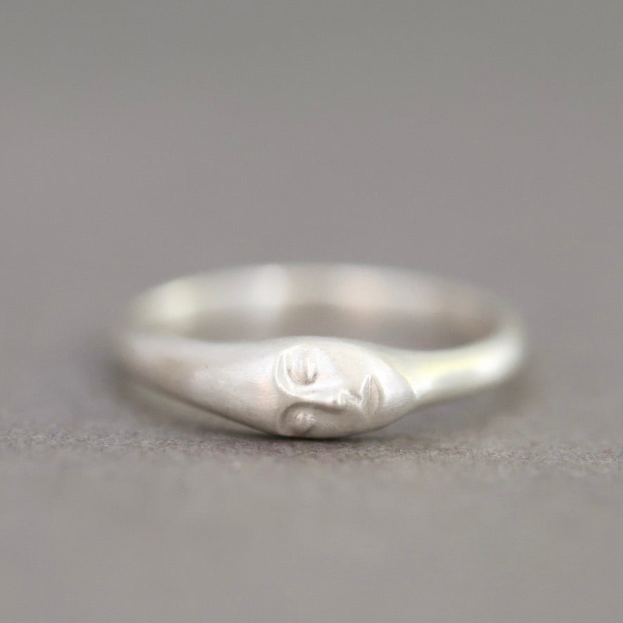 A sterling silver ring featuring a stylized, minimalist face carved into the band, set against a soft, neutral gray background.