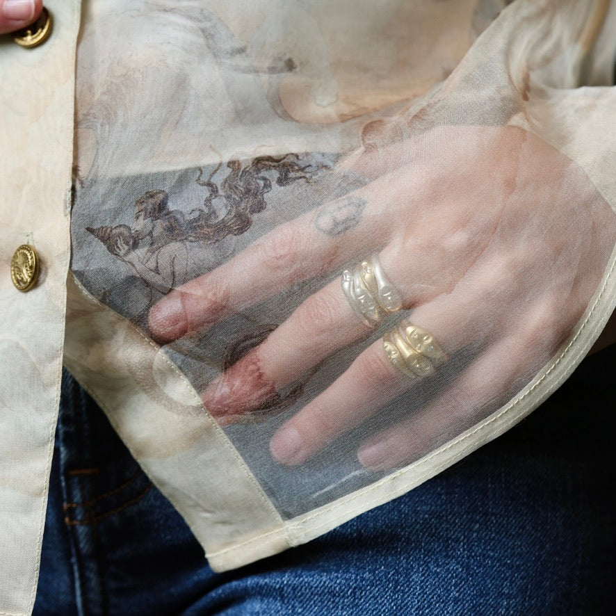 A close-up of a hand wearing two textured gold rings, resting on a sheer beige organza sleeve with a classical painting print, over a cream shirt and blue jeans.