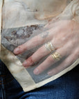 A close-up of a hand wearing two textured gold rings, resting on a sheer beige organza sleeve with a classical painting print, over a cream shirt and blue jeans.