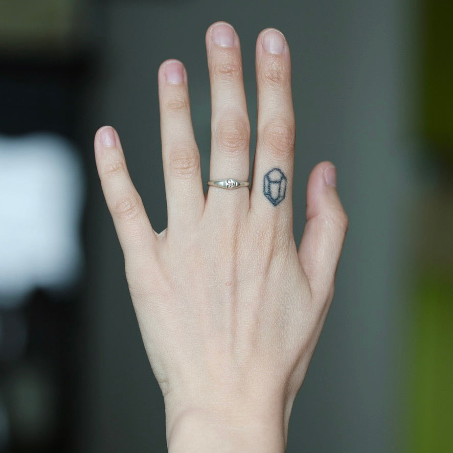 A close-up of a person's hand wearing a silver band ring with a small geometric gemstone, with a small black geometric tattoo on the ring finger.