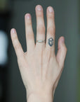 A close-up of a person's hand wearing a silver band ring with a small geometric gemstone, with a small black geometric tattoo on the ring finger.