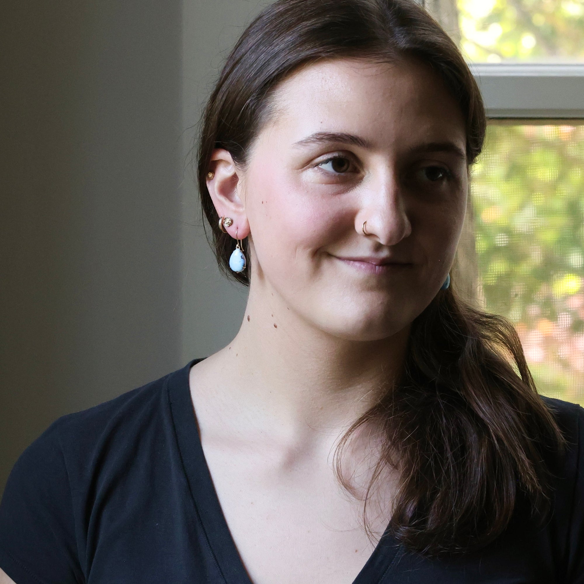 Woman with a nose ring and earrings standing indoors near a window with greenery outside.