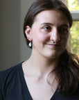 Woman with a nose ring and earrings standing indoors near a window with greenery outside.