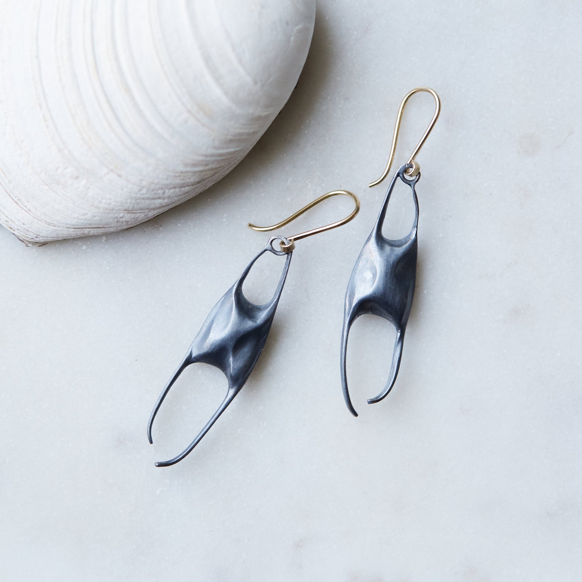 A pair of dangling earrings with dark metal sculptural drops and gold hook wires, displayed on a light marble surface next to a white clam shell.