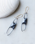 A pair of dangling earrings with dark metal sculptural drops and gold hook wires, displayed on a light marble surface next to a white clam shell.