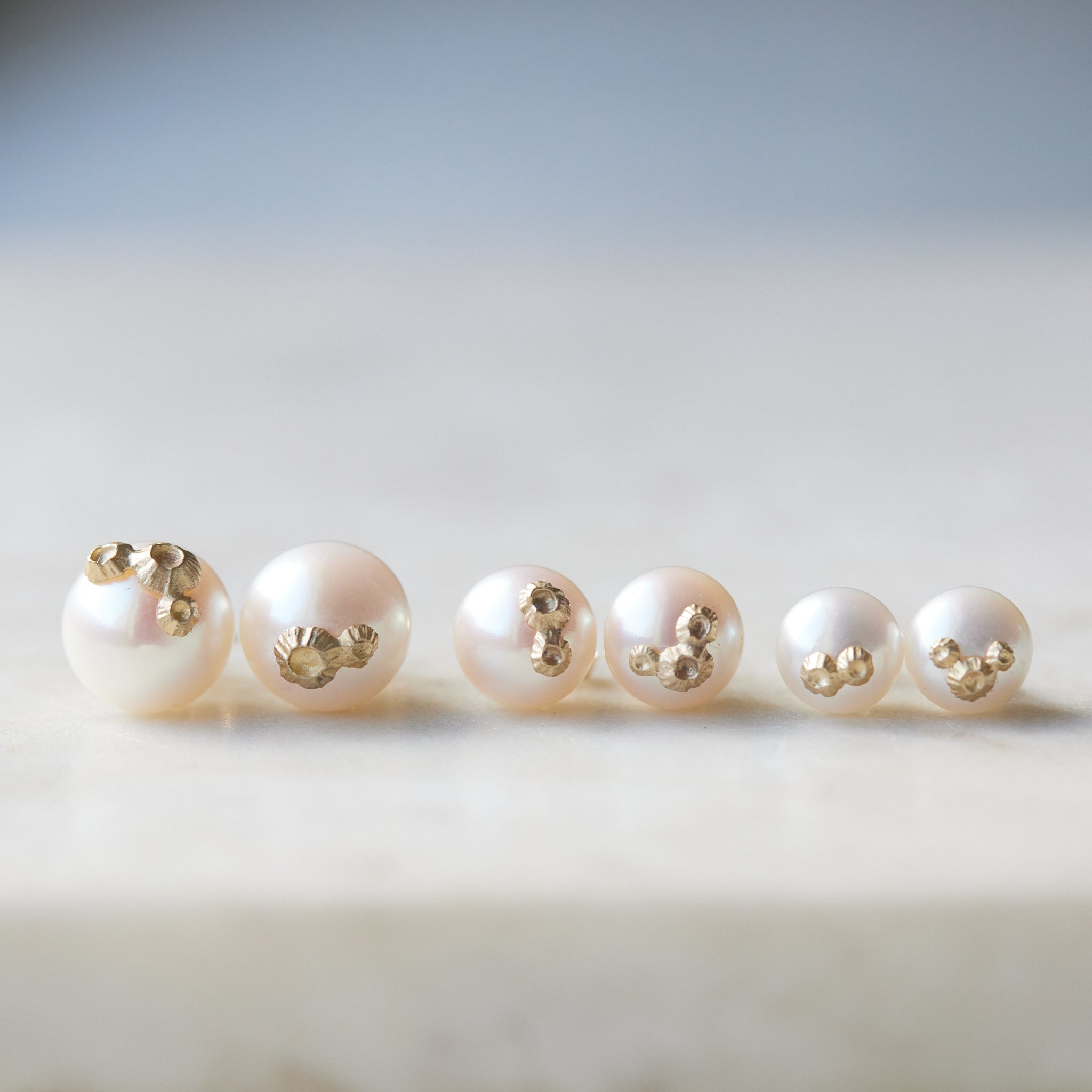 Three pairs of pearl studs in a row, photographed on white marble. From left to right, the large pearl studs with gold barnacles, medium pearl studs with gold barnacles, and little pearl studs with gold barnacles.