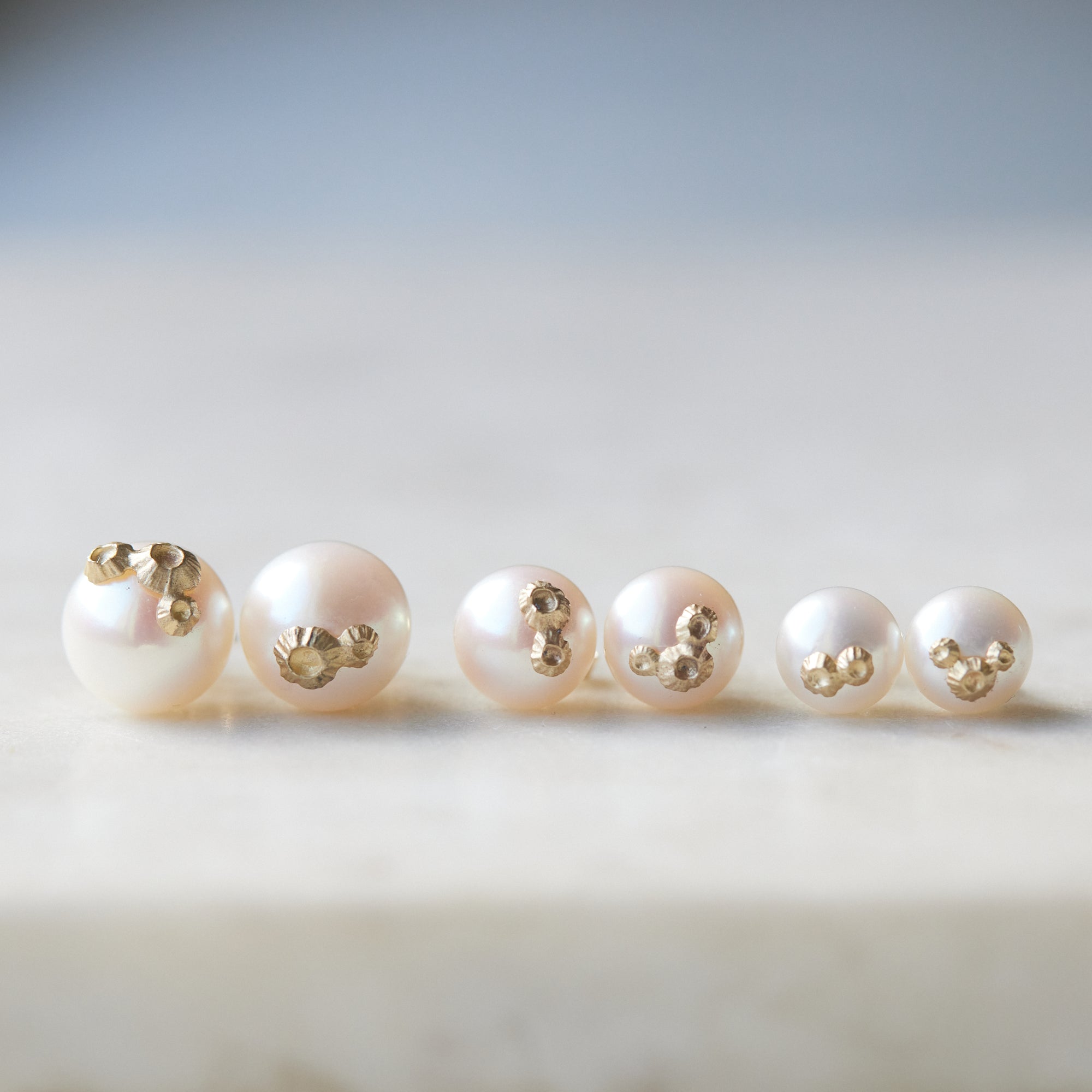 Three pairs of pearl studs in a row, photographed on white marble. From left to right, the large pearl studs with gold barnacles, medium pearl studs with gold barnacles, and little pearl studs with gold barnacles.