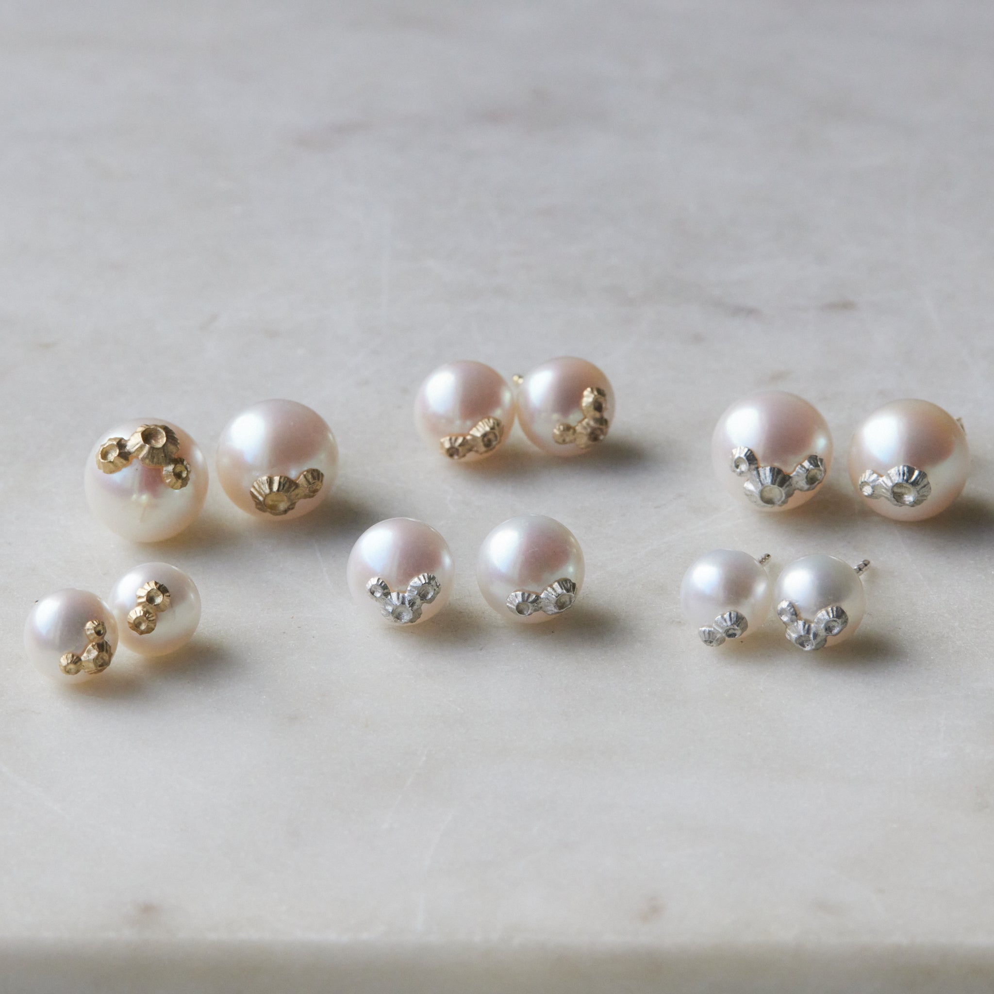 Six pairs of pearl studs in three sizes and two metals, photographed on white marble. The back row shows, left to right, the large pearl studs with gold barnacles, medium pearl studs with gold barnacles, and large pearl studs with silver barnacles. The front row shows, left to right, the little pearl studs with gold barnacles, the medium pearl studs with silver barnacles, and the little pearl studs with silver barnacles. 