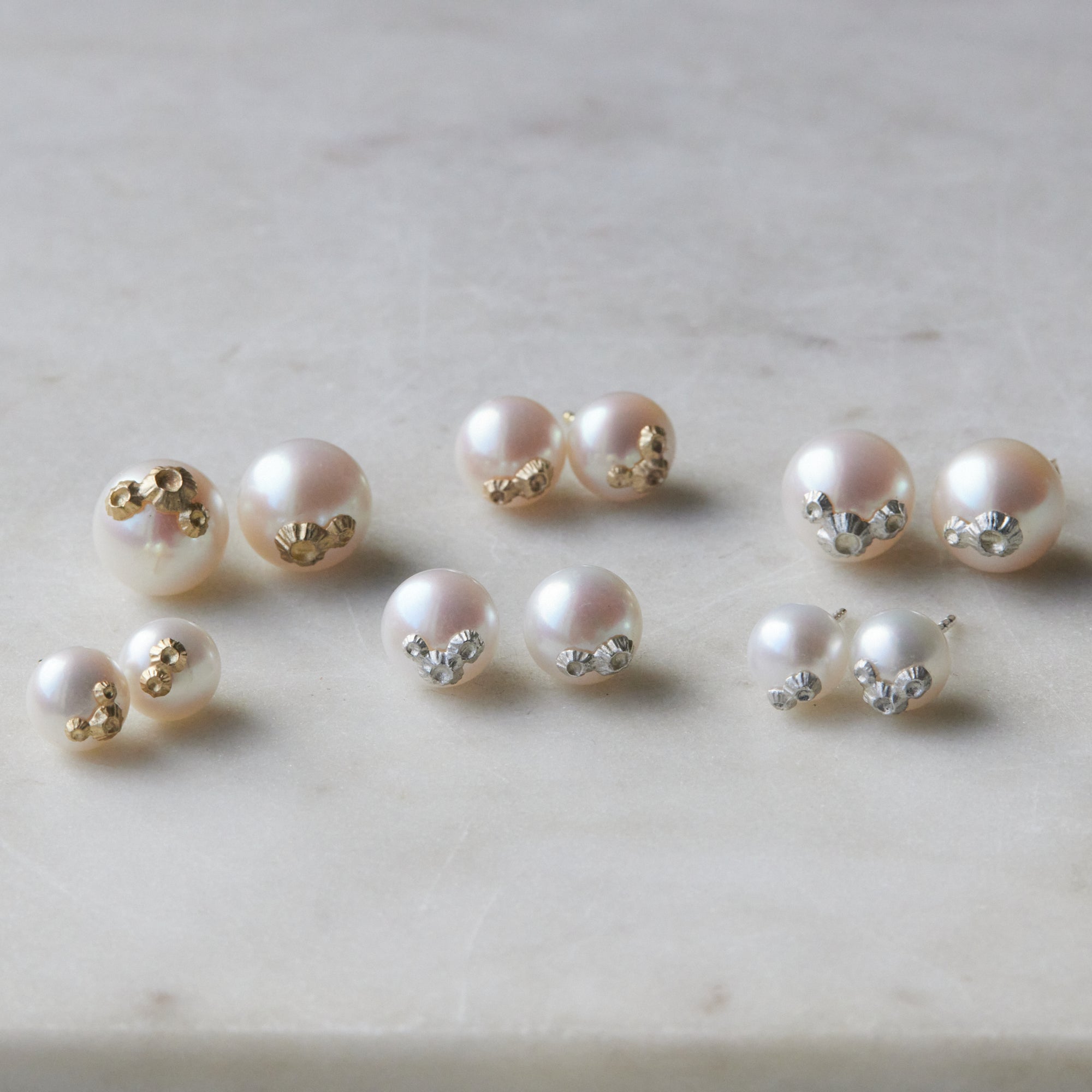 Six pairs of pearl studs in three sizes and two metals, photographed on white marble. The back row shows, left to right, the large pearl studs with gold barnacles, medium pearl studs with gold barnacles, and large pearl studs with silver barnacles. The front row shows, left to right, the little pearl studs with gold barnacles, the medium pearl studs with silver barnacles, and the little pearl studs with silver barnacles. 