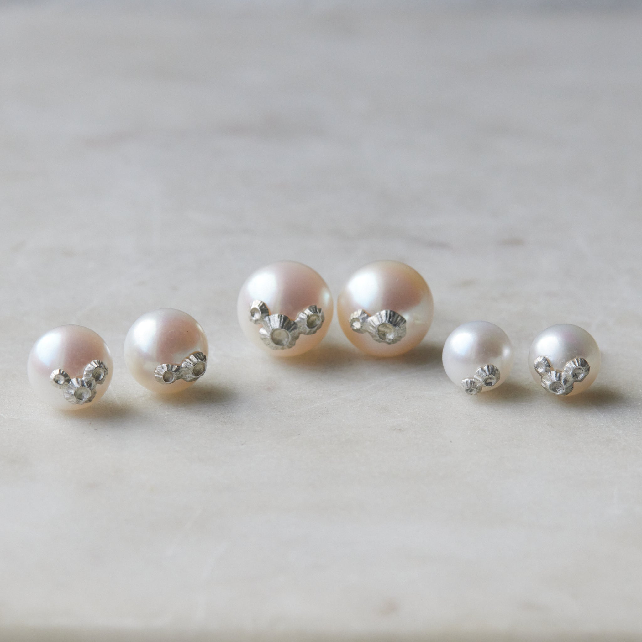 Three pairs of pearl studs in a row, photographed on white marble. From left to right, the medium pearl studs with silver barnacles, the large pearl studs with silver barnacles, and the little pearl studs with silver barnacles.