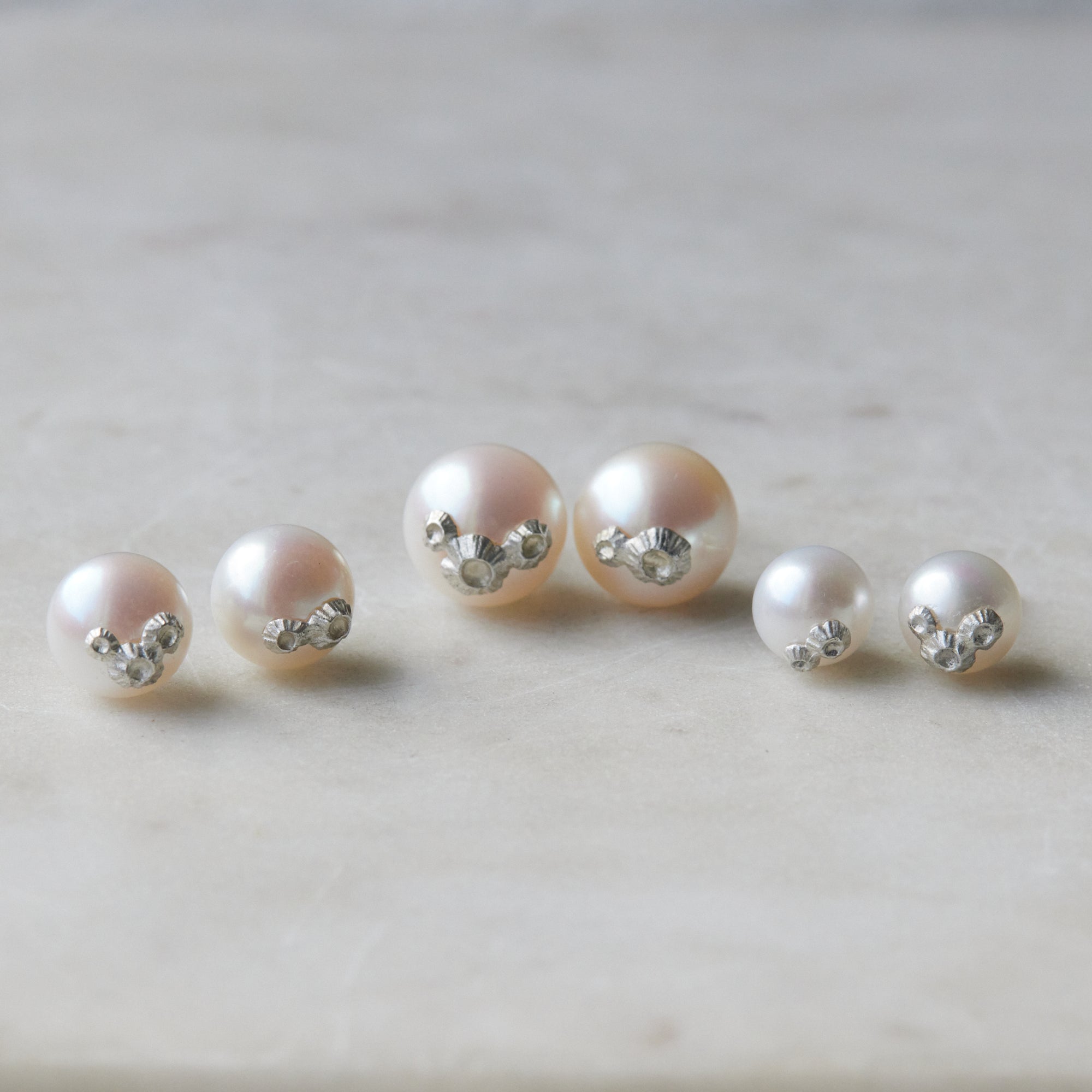 Three pairs of pearl studs in a row, photographed on white marble. From left to right, the medium pearl studs with silver barnacles, the large pearl studs with silver barnacles, and the little pearl studs with silver barnacles.