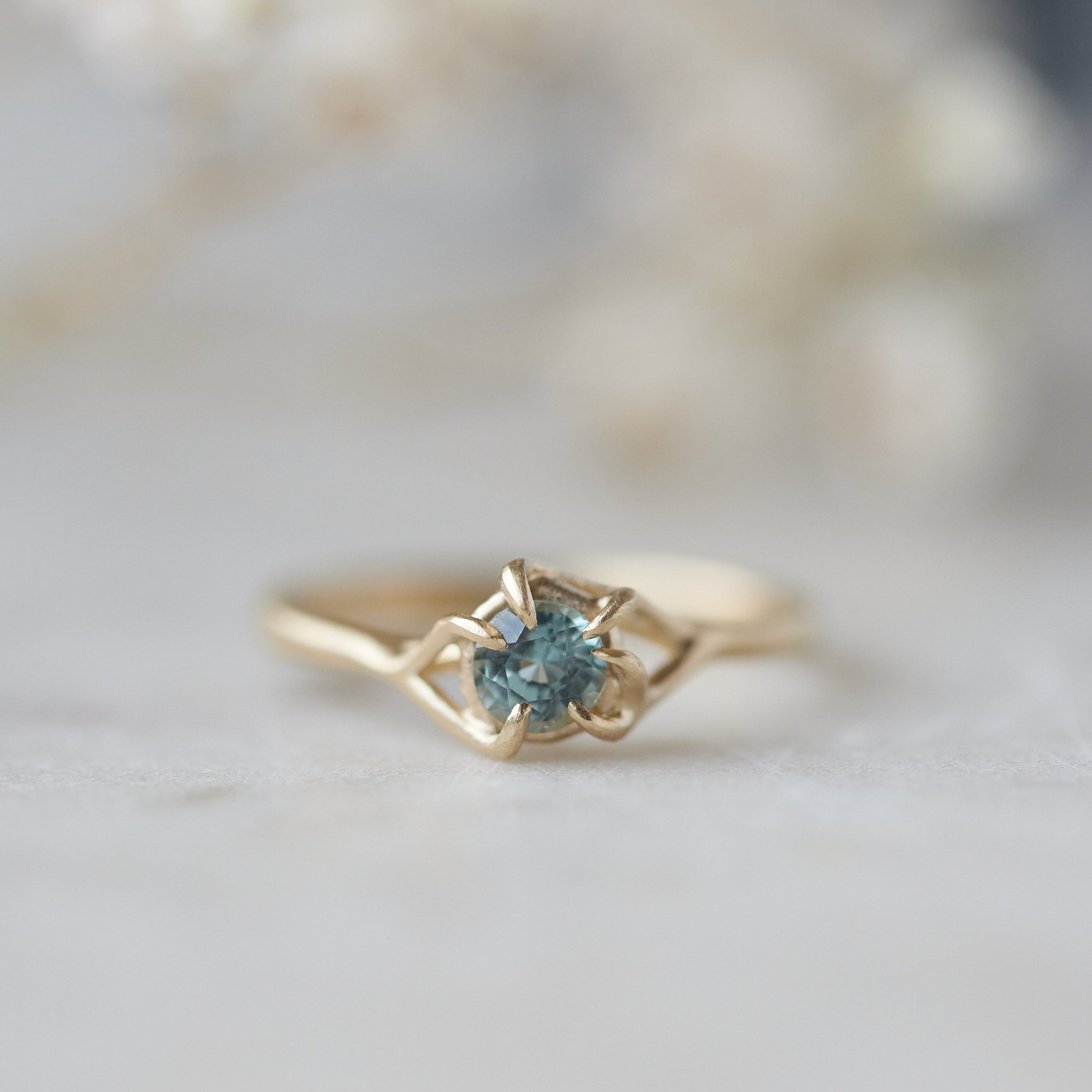 a round crystal blue montana sapphire is wrapped in gold branches. photographed on a white marble slab.