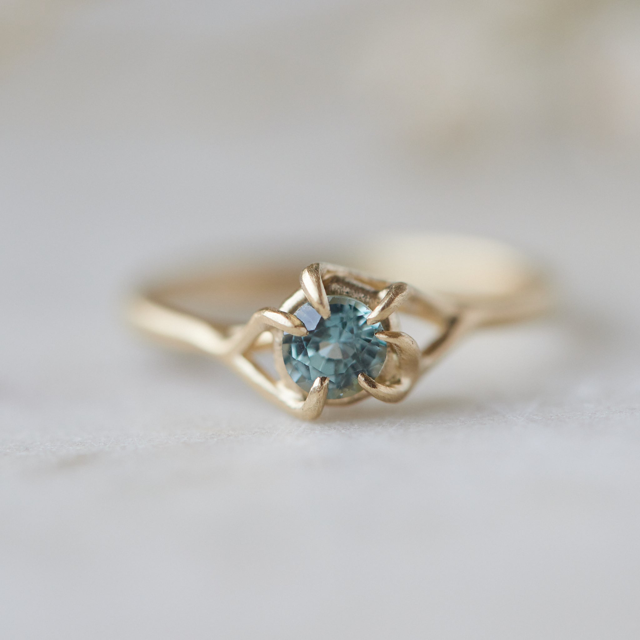 a round crystal blue montana sapphire is wrapped in gold branches. photographed on a white marble slab.